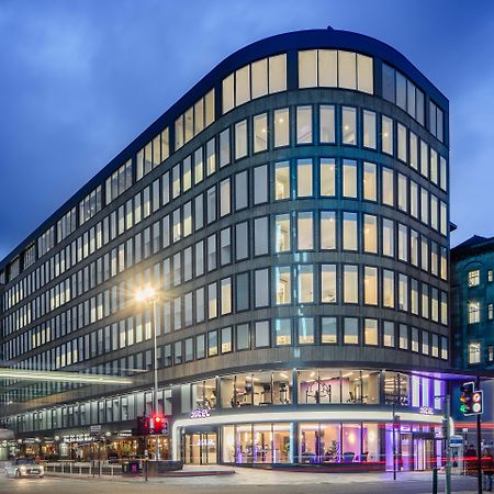 Yotel غلاسغو المظهر الخارجي الصورة The photo shows a modern office building with a distinctive curved shape and a facade made of large glass windows. It is illuminated against a twilight sky, giving it a contemporary appearance. The ground floor features large glass entrance areas, li