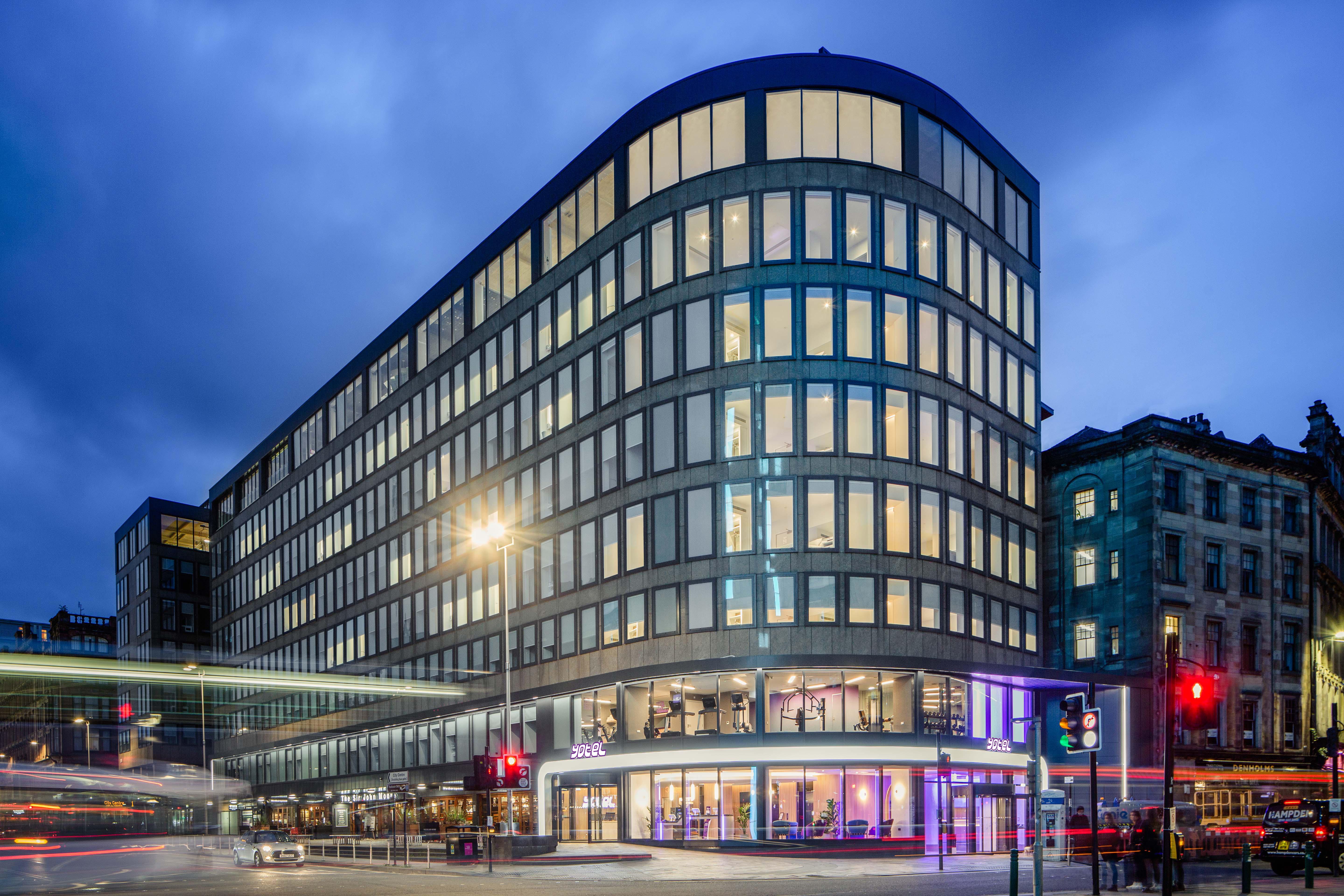 Yotel غلاسغو المظهر الخارجي الصورة The photo shows a modern office building with a distinctive curved shape and a facade made of large glass windows. It is illuminated against a twilight sky, giving it a contemporary appearance. The ground floor features large glass entrance areas, li